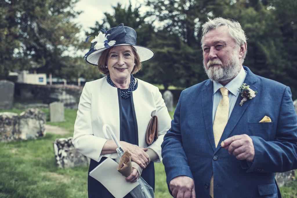 Woman in white dress and hat and man with grey beard in blue suit at an event
